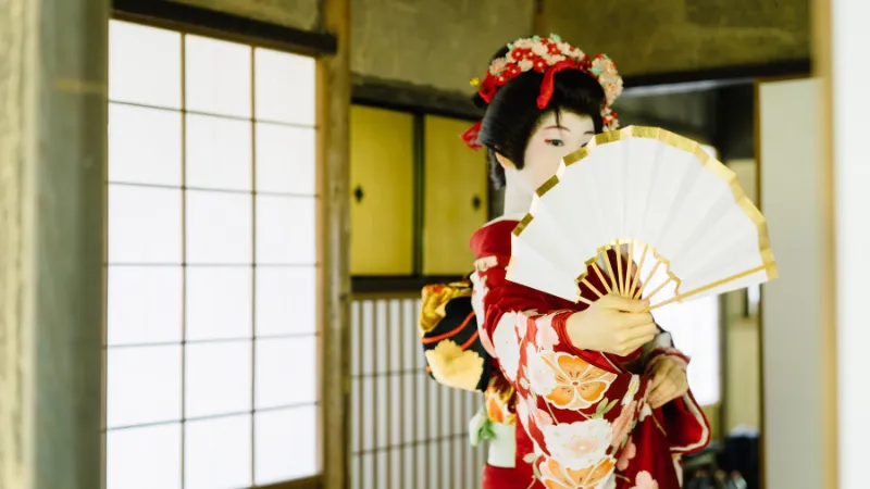 障子のある部屋で、扇子で半分顔を隠した日本髪の着物女性