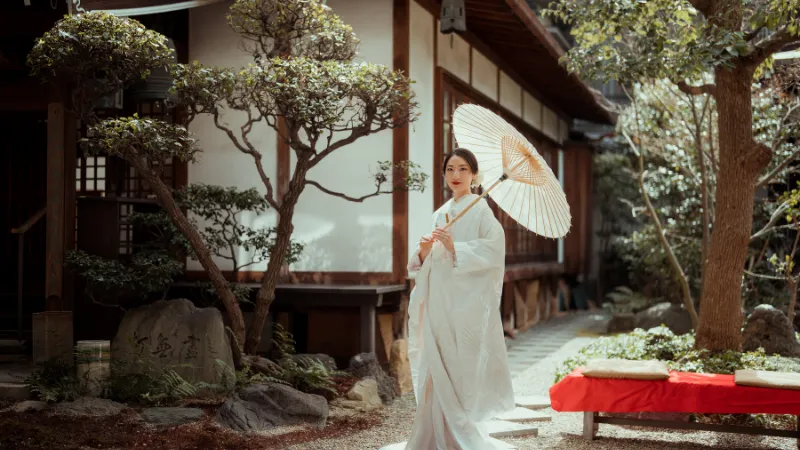 白い蛇の目傘を差し、白いお引きずりの着物姿の女性が日本庭園に佇んでいる