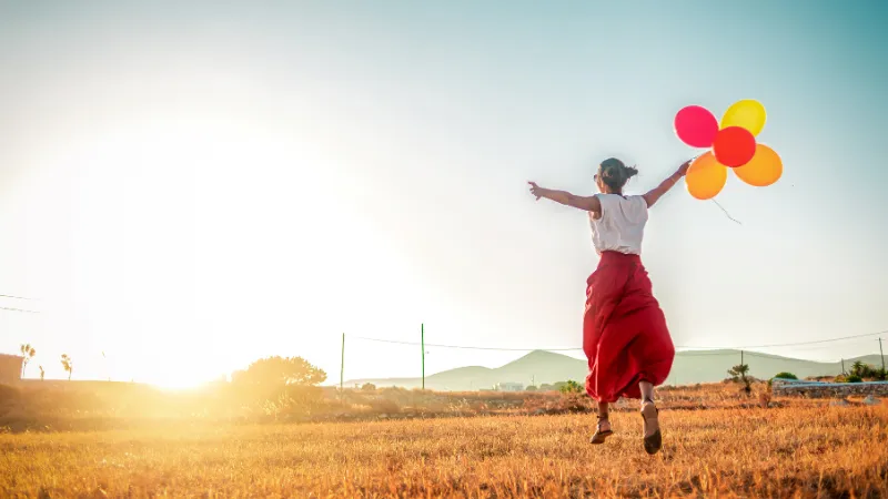 日の当たる草原の上で風船を持って手を広げてジャンプする女性