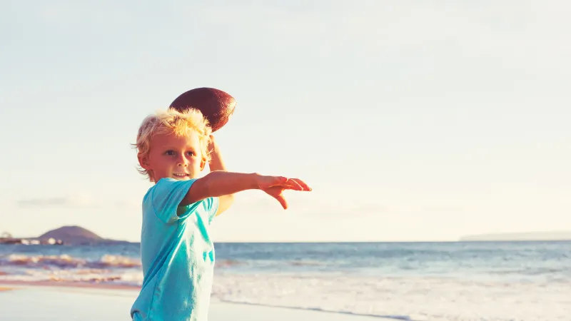 太陽の光が降り注ぐ海岸で、ボールを投げようとしている金髪の幼い男の子